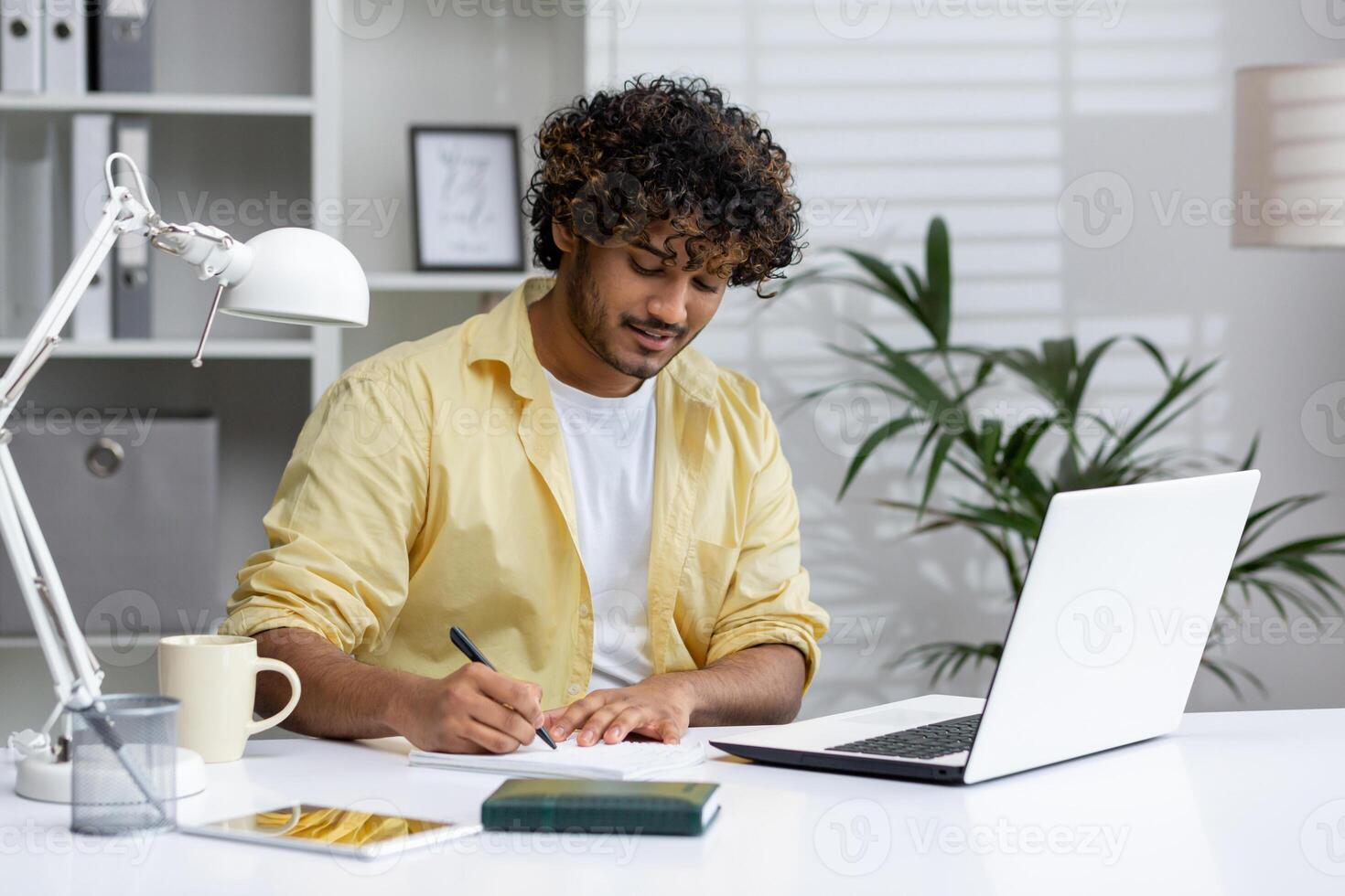 Man working from home office, writing in notebook, using laptop, and enjoying a coffee. Modern workspace with plants and natural light. photo
