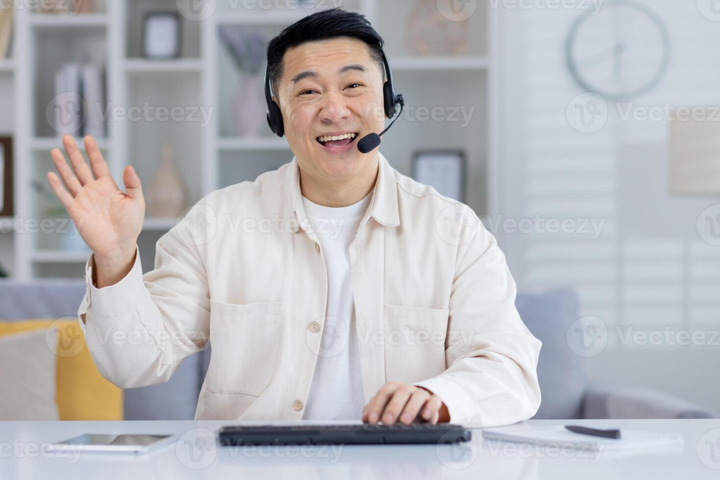 Cheerful Asian man waving during a conference at home. He sits in a well-lit room, showcasing a professional yet relaxed atmosphere. photo