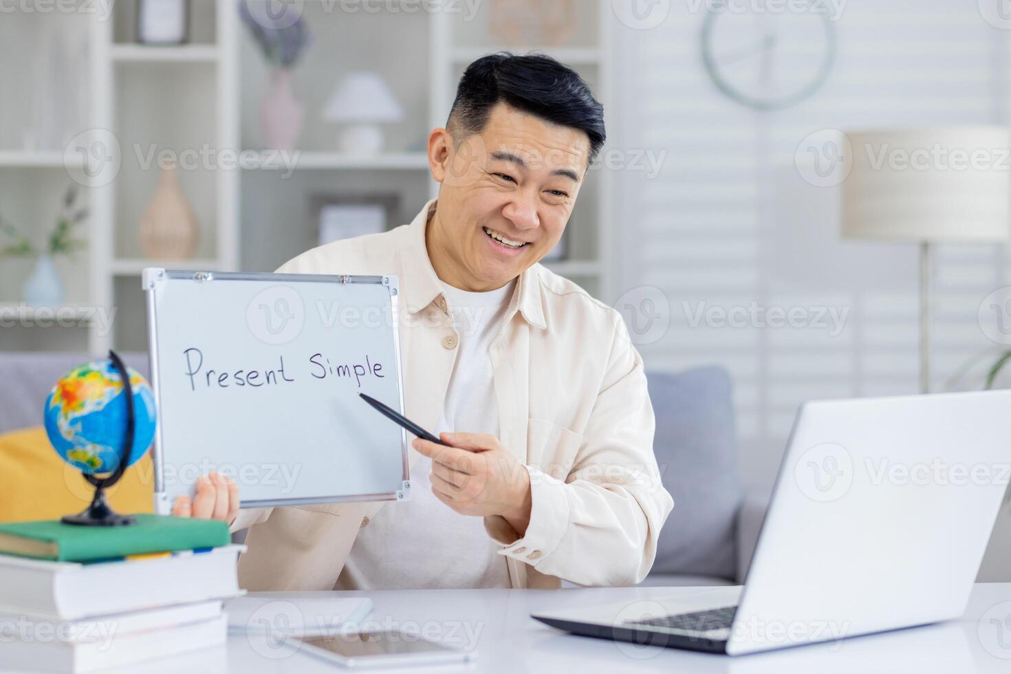 Smiling online language teacher explaining present simple tense during a virtual lesson. Man holding a whiteboard and pointing with a pen while teaching via laptop. photo
