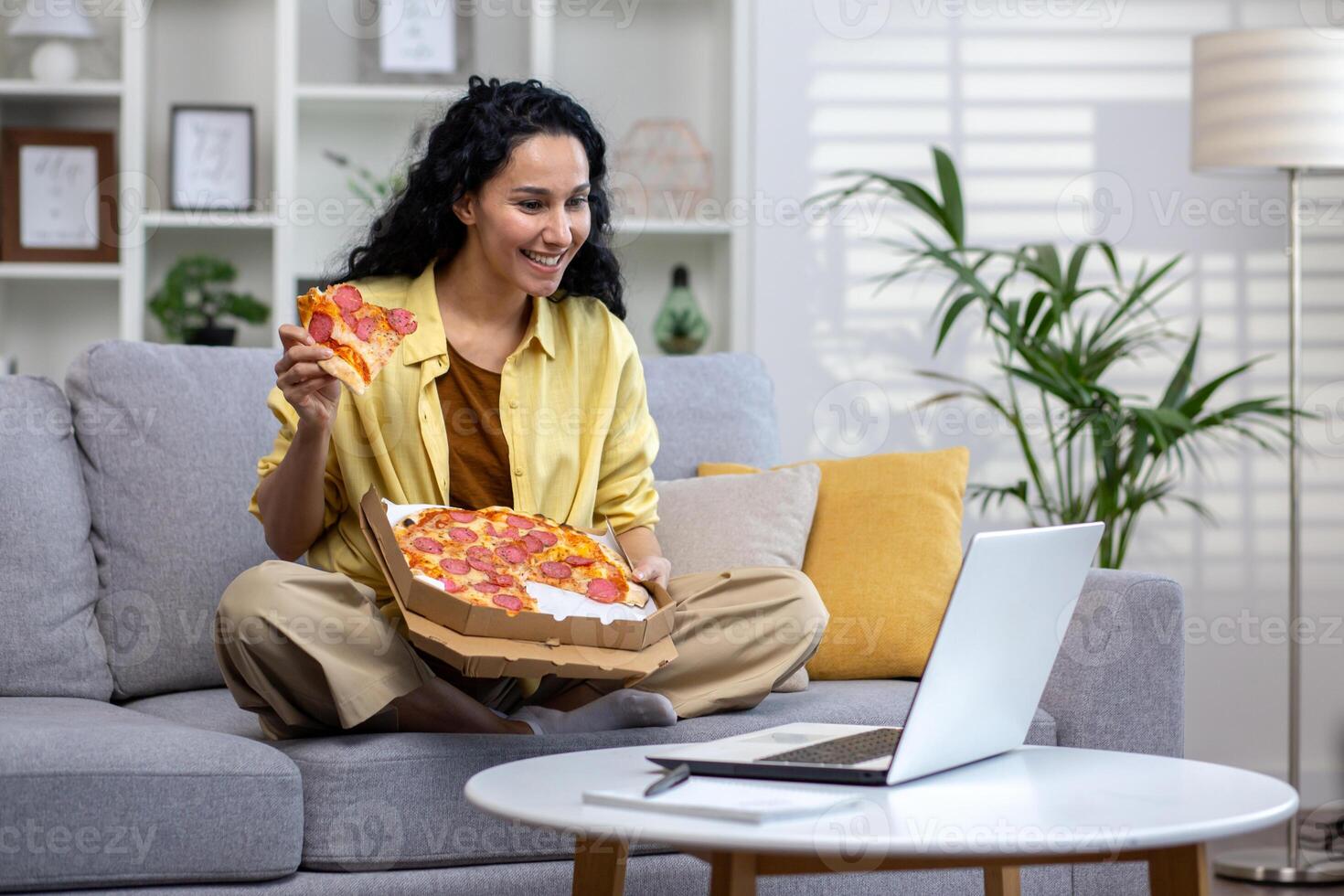 Joyful and happy woman eating pizza alone at home, Hispanic woman satisfied with timely food delivery, using laptop to watch online stream, sitting on sofa in living room at home. photo