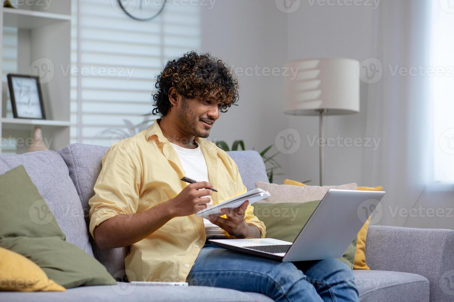 A young adult sits comfortably on a sofa, working on a laptop while taking notes in a notebook, depicting a productive work-from-home setting. photo