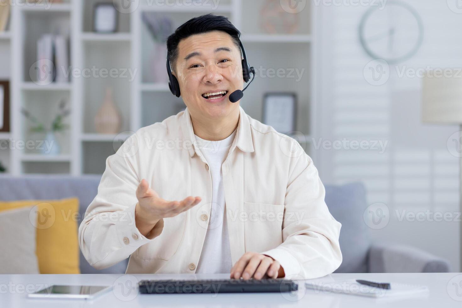 Cheerful Asian male in a beige shirt using a headset in a home office, looking at camera with a smile, possibly in a customer service call. photo