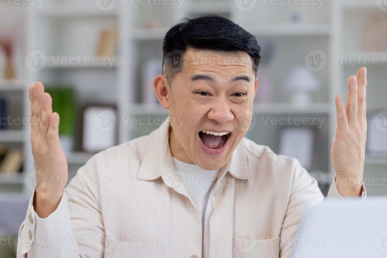 A middle-aged Asian man shows great excitement and joy while working at his computer in a modern home office setting. His exhilarated expression suggests a moment of triumph. photo