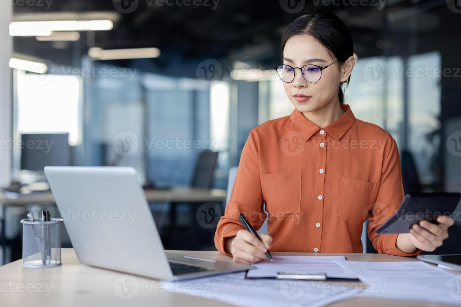 Serious businesswoman in glasses working at her office desk with a laptop, documents, and a tablet, focusing on her tasks. photo