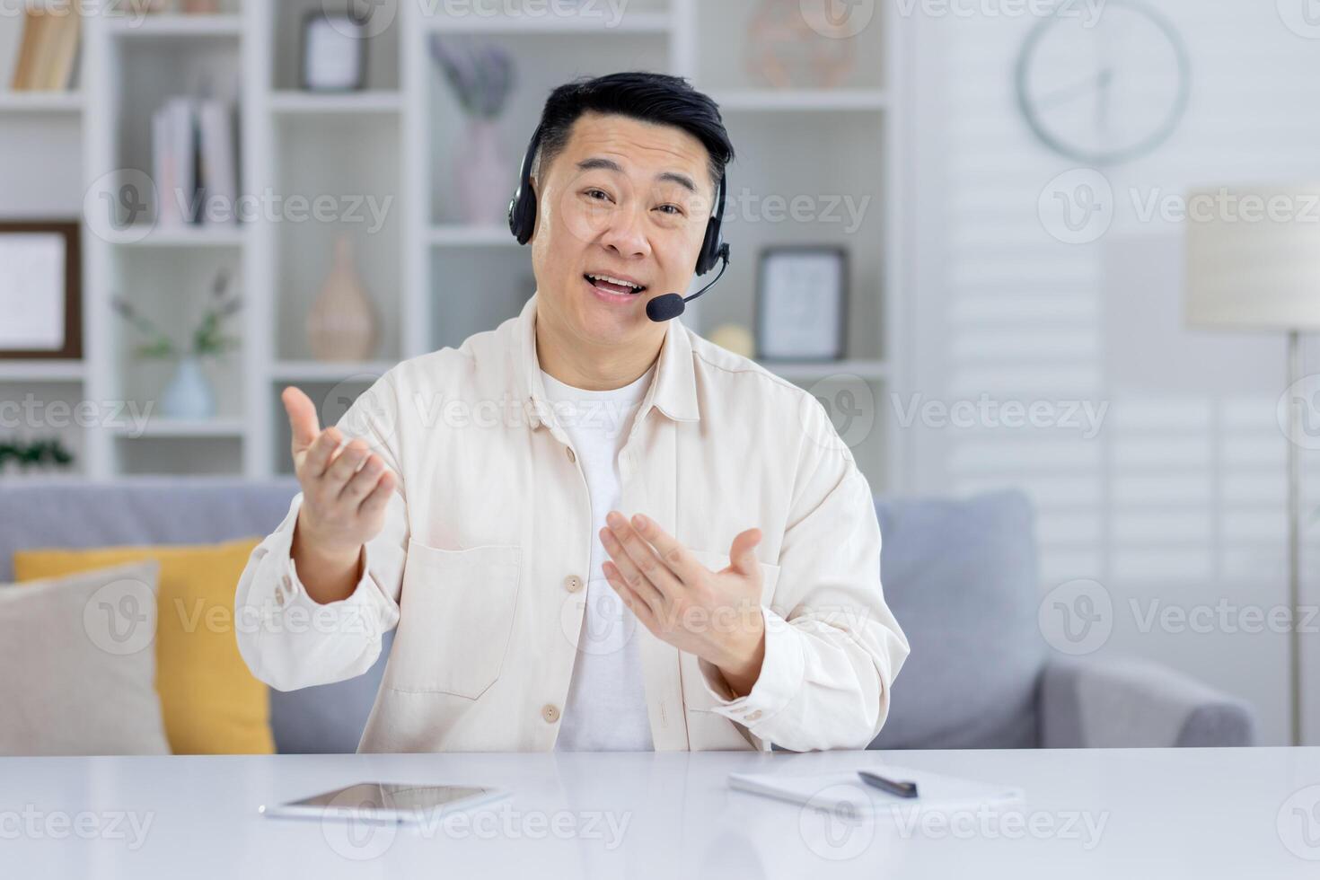 Enthusiastic Asian man wearing a headset engaged in a lively conversation, gesturing with hands in a bright home office setting. photo
