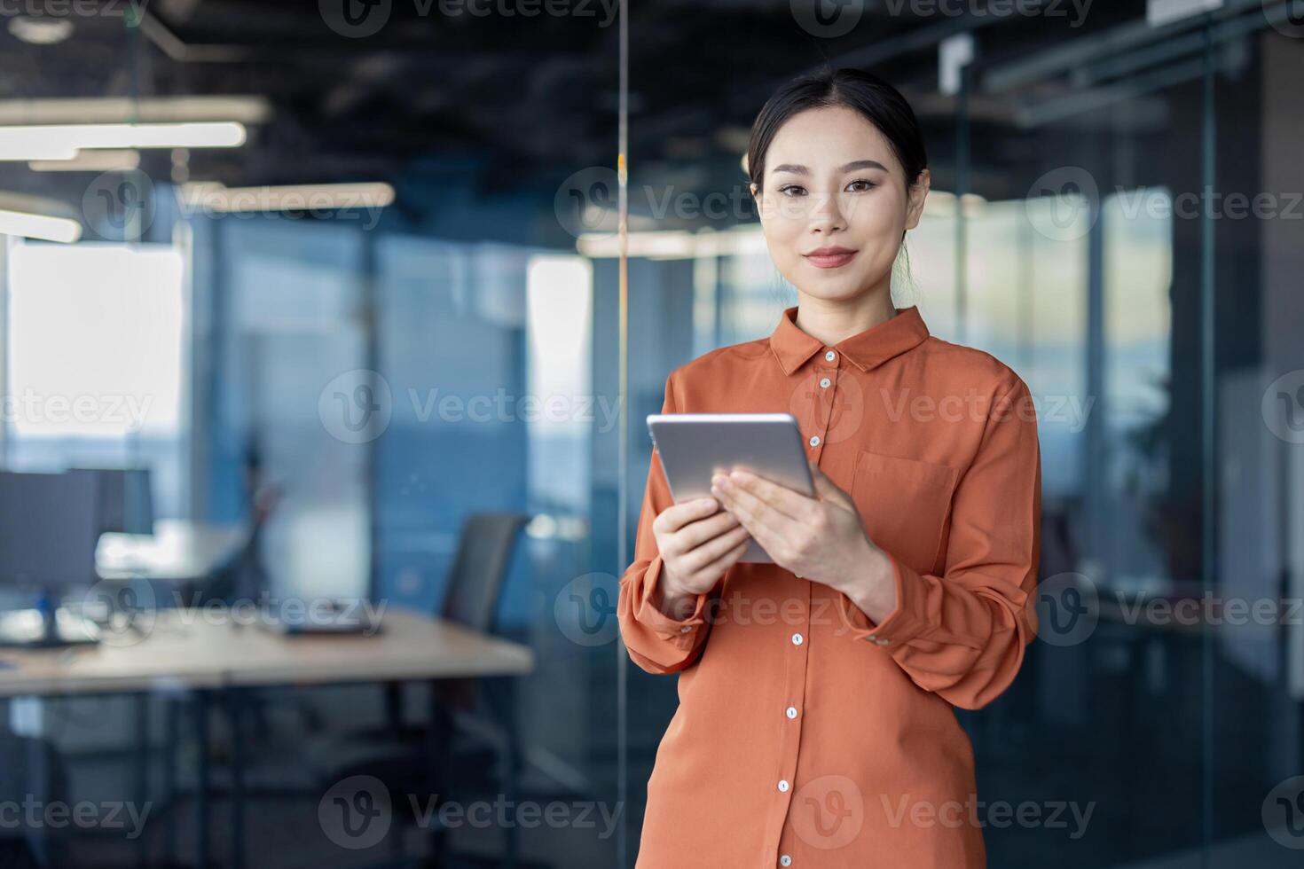 A professional Asian businesswoman looks directly at the camera while holding a digital tablet in a well-lit, modern office environment, symbolizing tech-savvy business operations. photo