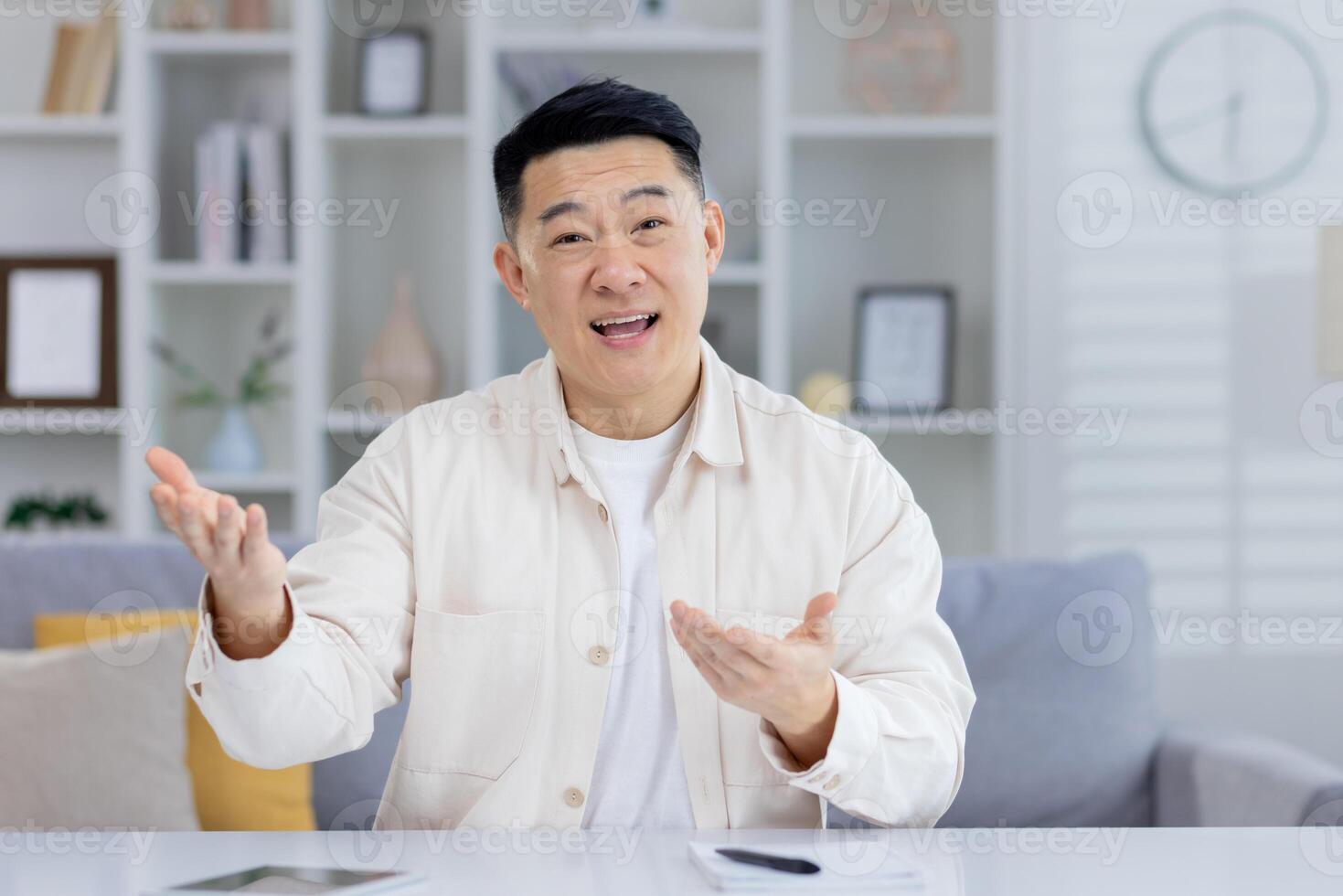 Cheerful Asian man engaging in a casual conversation at home, looking at the camera and talking with expressive hand gestures in a modern living room setting. photo