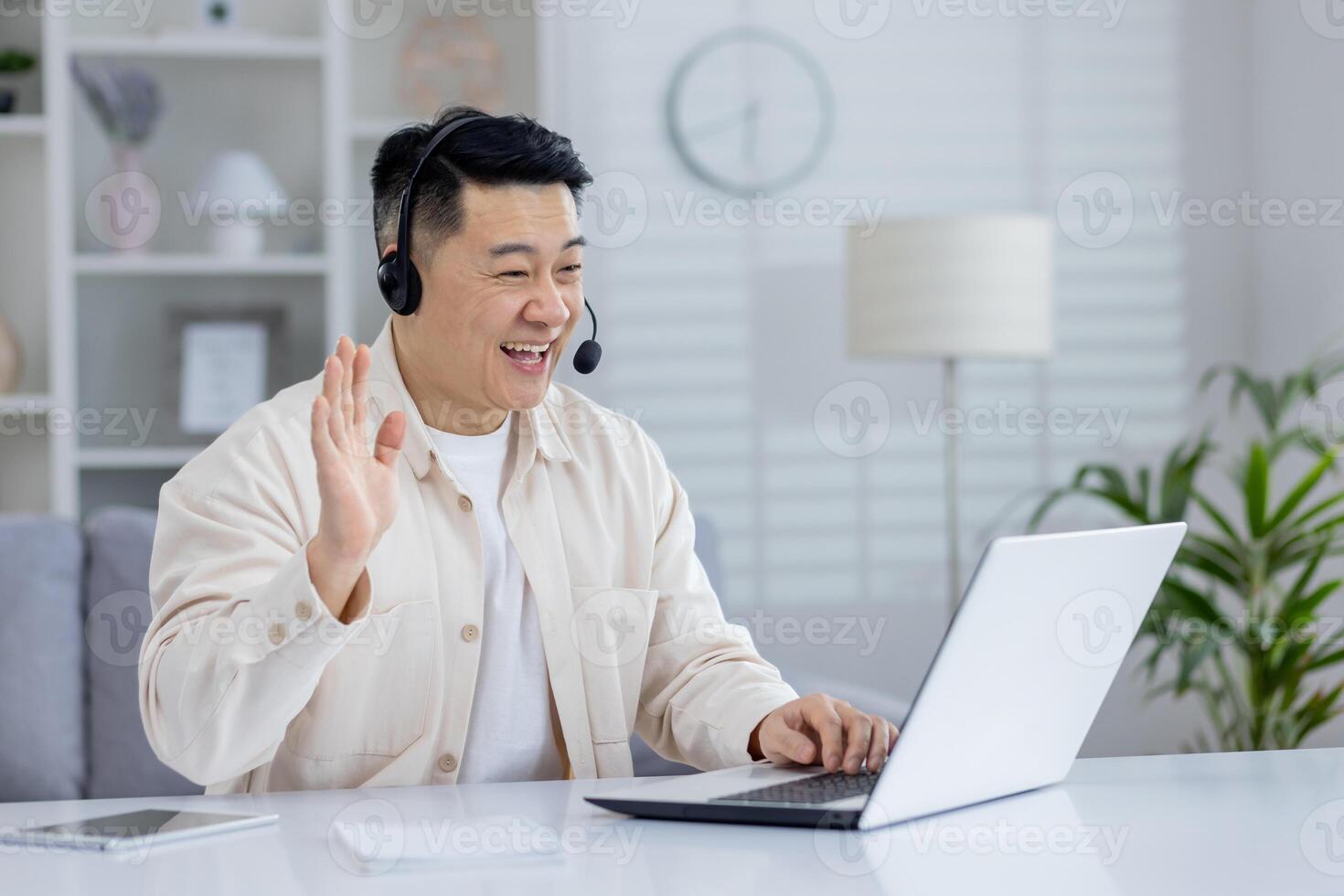 Cheerful Asian adult male using a laptop and headset at a bright home office setup, waving while on a call. photo