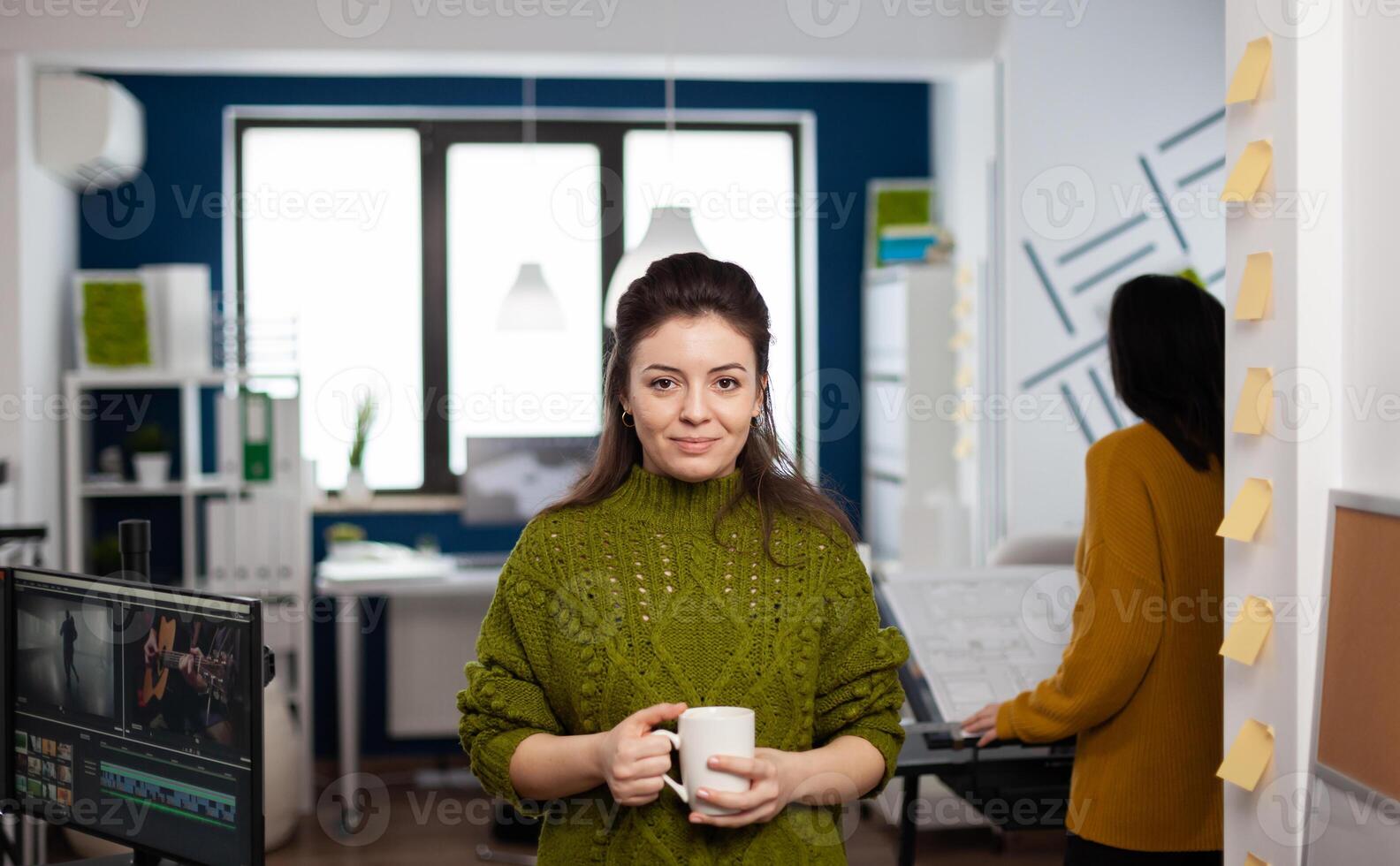 Portrait of creative designer smiling at camera holding a cup of coffee standing in start up agency office. Woman working in multimedia studio production editing in workplace with diverse team. photo