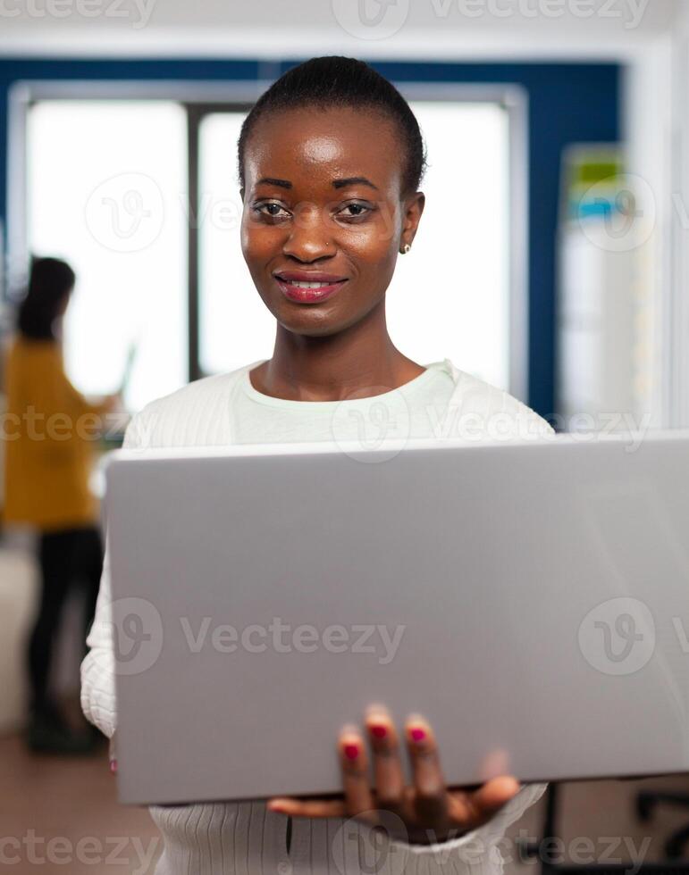African videographer smiling at camera holding at laptop standing in creative agency office. Videographer working in multimedia studio production editing in modern workplace. photo