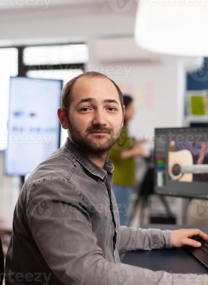 Man editor looking at camera, working with footage and sound on computer with two displays. Videographer editing movie smiling while processing film montage sitting in startup office. photo