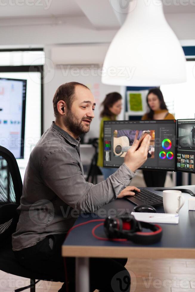Videographer discussing online with customer holding smartphone editing film footage sitting in creative agency. Man editor processing movie montage in multimedia company with post production software photo