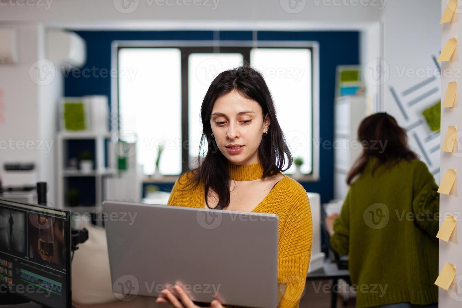 Focused entrepreneur standing in creative agency office holding laptop and typing project informations. Photographer graphic editor working in start up digital multimedia company, using software app photo