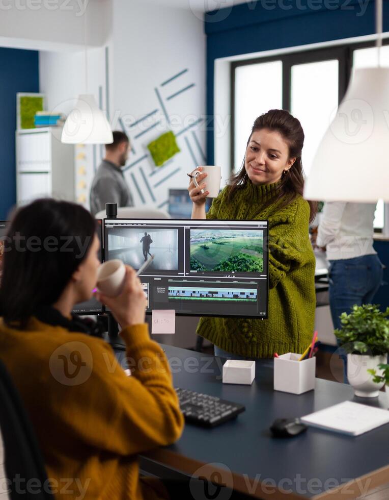 Team of videographers talking about project while drinking coffee working in editing app at pc sitting in digital multimedia company. Creative women having break while prosessing film montage photo