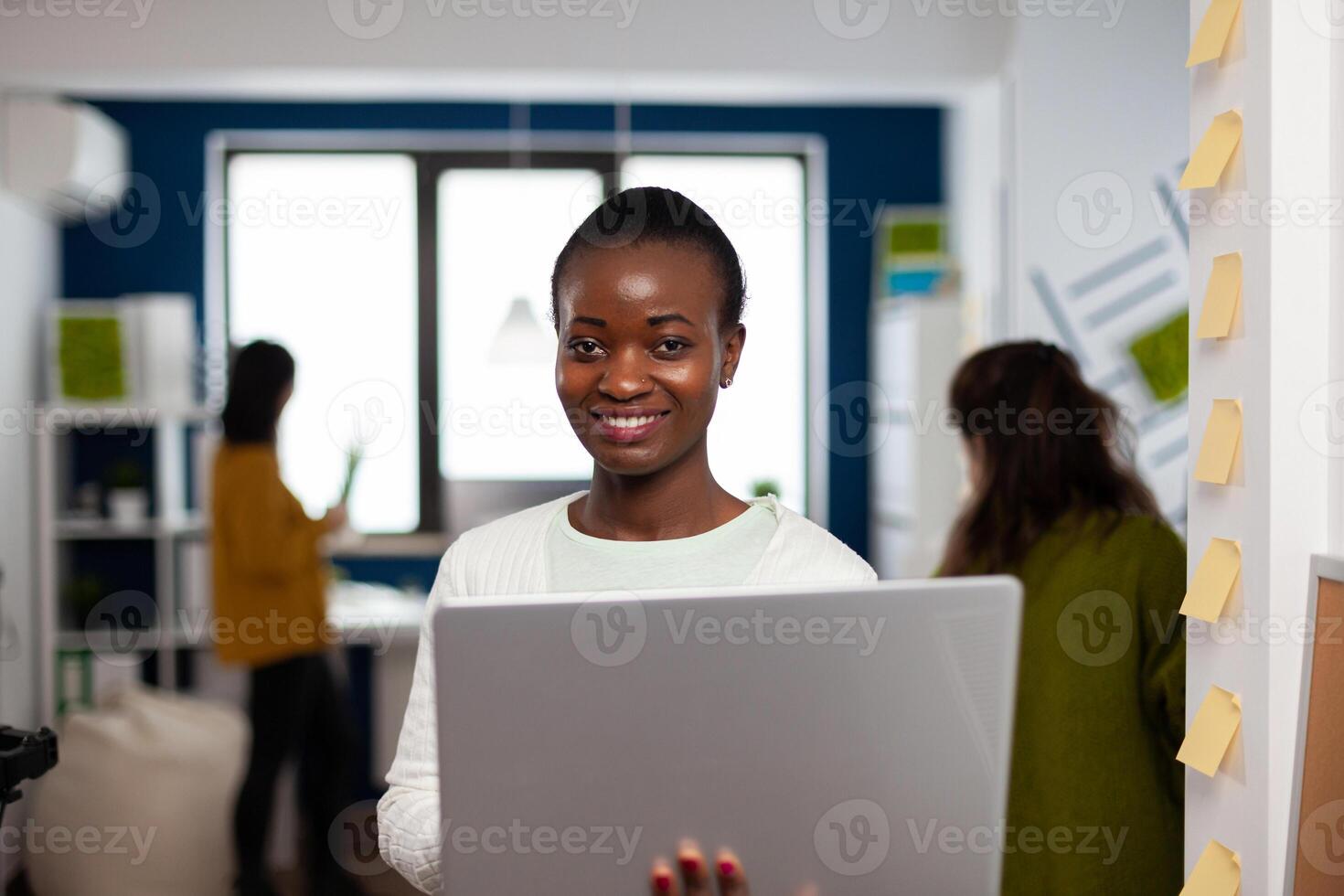 Close up of african woman looking at camera smiling standing in start up creative agency office holding laptop, typing on it. Black videographer editing project using post production software photo