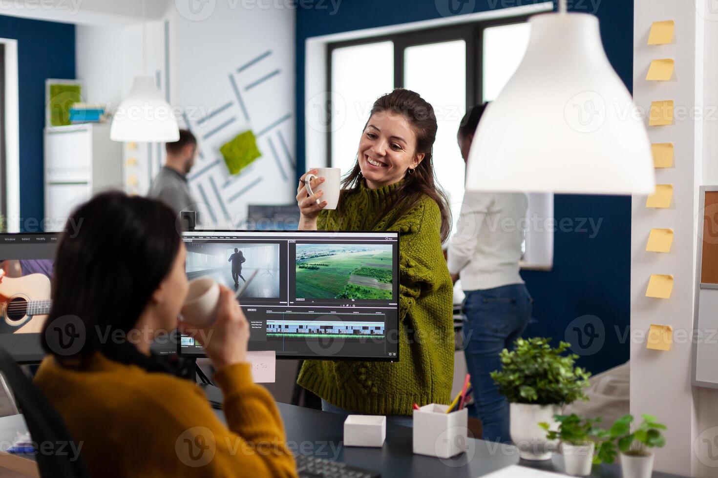 Videographers drinking coffee and talking during work break sitting at desk in creative agency office. editor and sound engineer working at project, editing film footage in software app photo