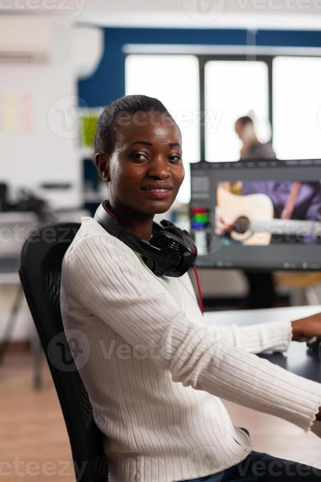 African editor looking at camera smiling editing project in post production software working in creative studio office. Videographer editing audio film montage on professional computer photo
