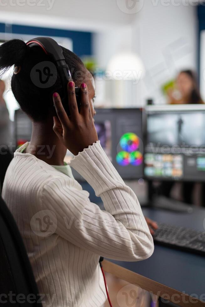 Black editor working with footage on pc with two monitors, putting on headphones. African videographer editing movie using post production software working in creative start up agency office. photo