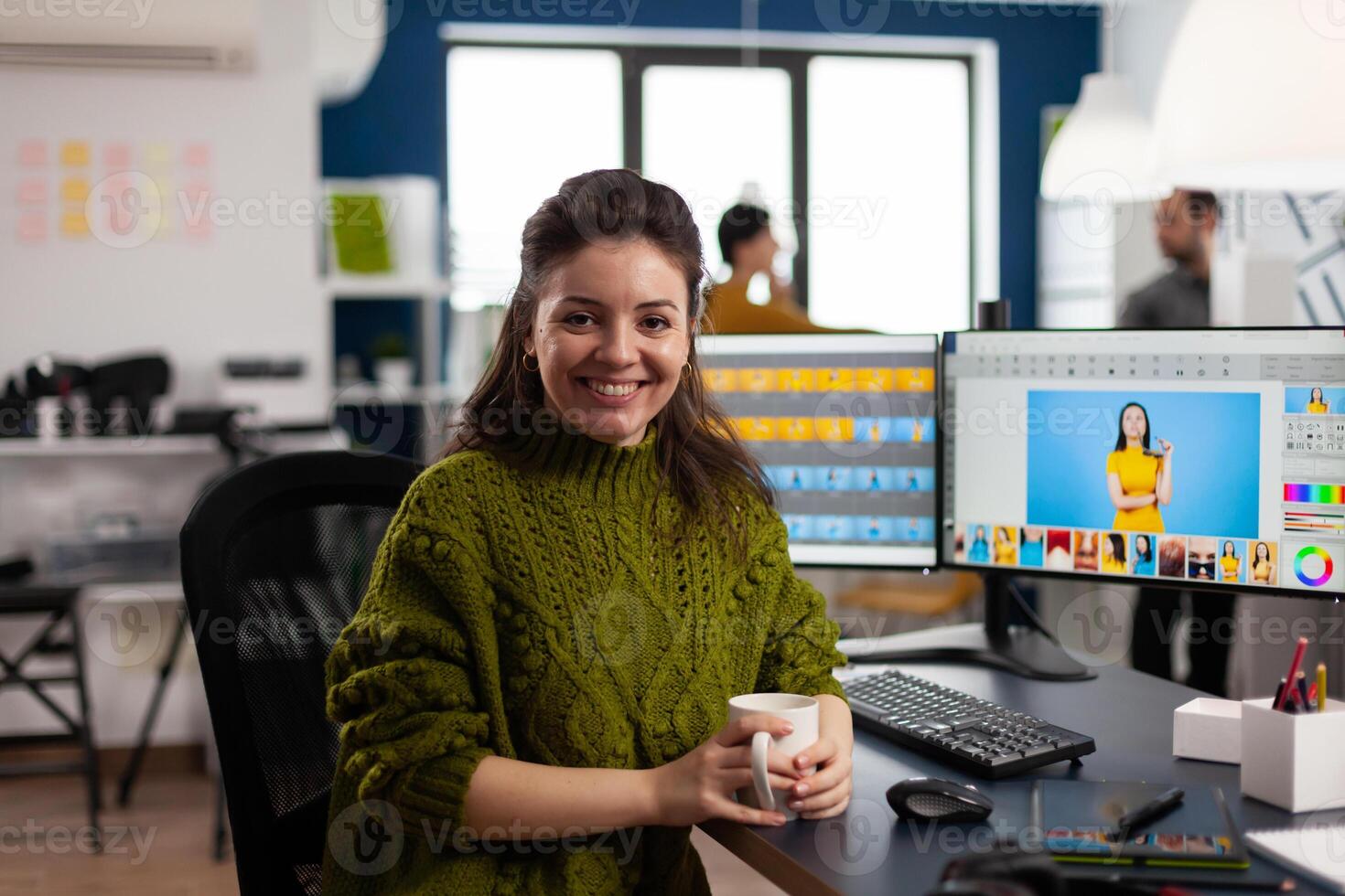 Woman retoucher looking at camera smiling sitting in creative media agency retouching client photos on PC with two displays. Photographer graphic editor working in start up office with digital assets