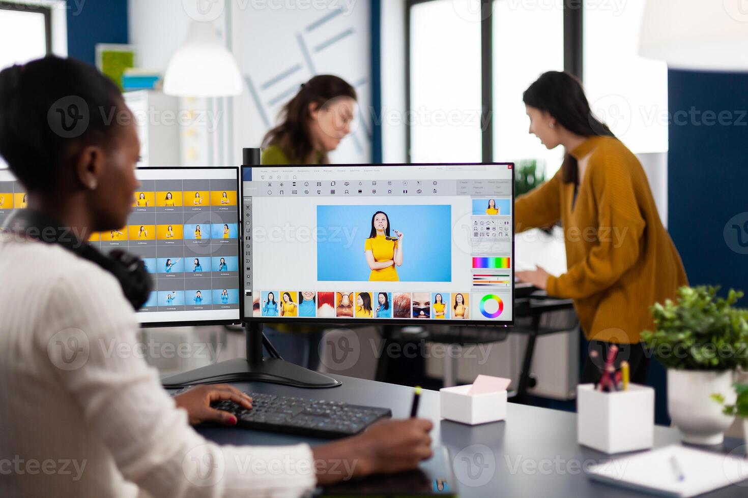African woman retoucher sitting in modern creative photo studio working on computer after photoshoot, retouching photographs of model in image editing software with stylus pencil, color greading app.