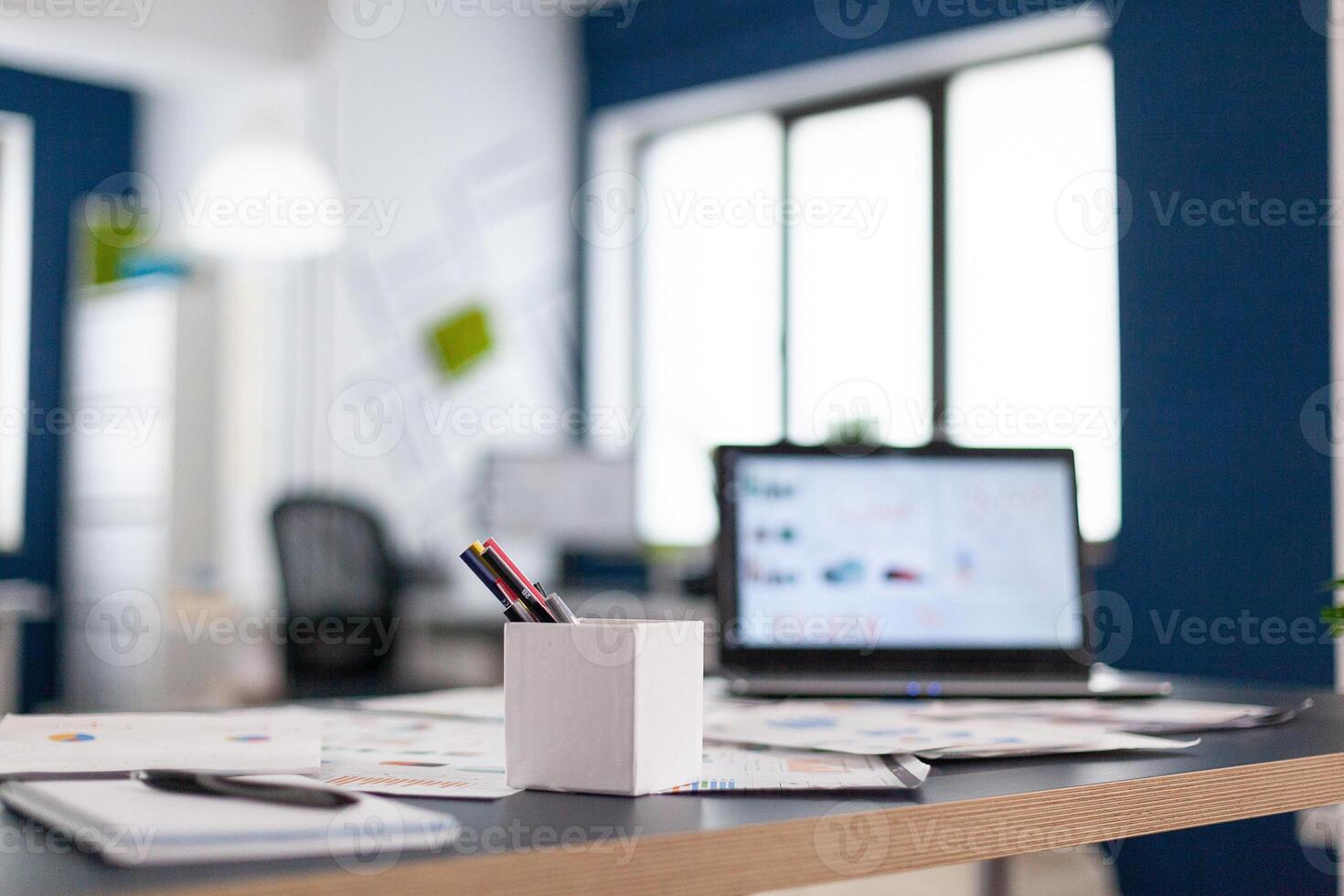 Laptop with company financial charts on office desk. Brainstorm area in business center with nobody in it, shot of empty room with modern furniture and blue wall. photo