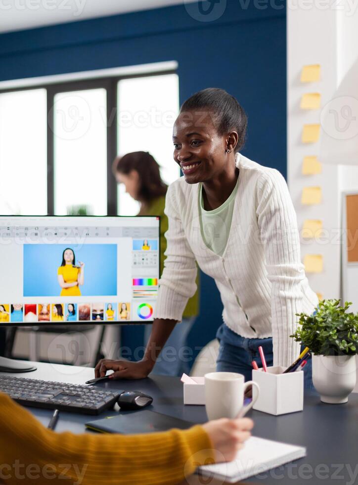 Satisfied african photo editor discussing with colleague how retouching image, working on PC. Woman retoucher editing assets in pro software app with graphic tablet sitting in creative agency