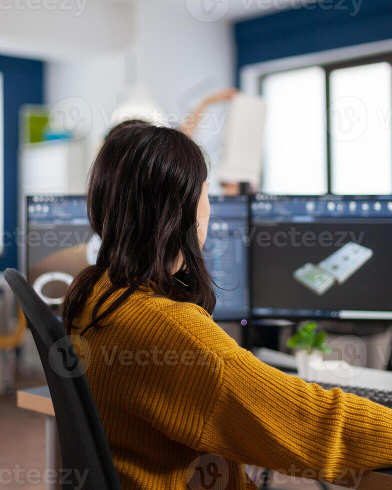 Young woman industrial engineer working at pc with two monitors, screens showing CAD software with 3D prototype of gears metalic mechanical piece. Employee working in creative agency office photo