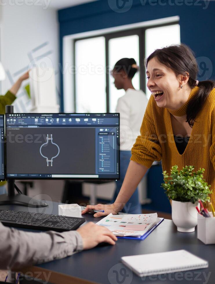 Excited mechanical engineers laughing and discussing about manufacturing new project, taking break. Man technician working at pc, screen shows 3D engine made with CAD software, using computer photo