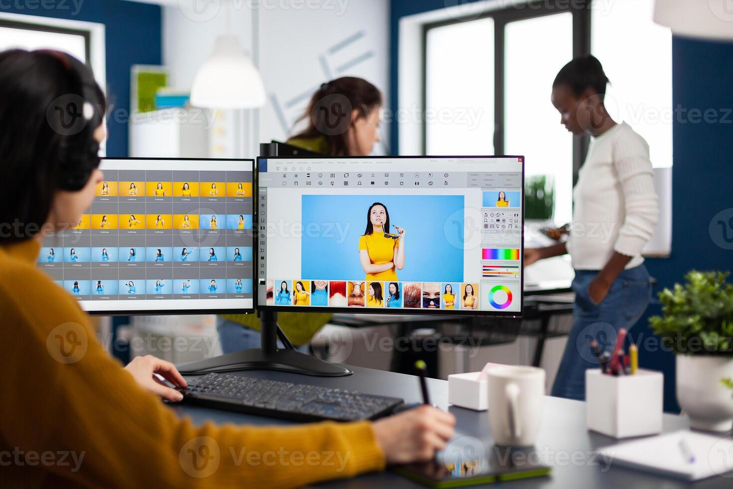 Woman retoucher with headset retouching photos using stylus pen drawing on graphic tablet, working on PC with two displays. Photo editor working in software app sitting in creative agency office