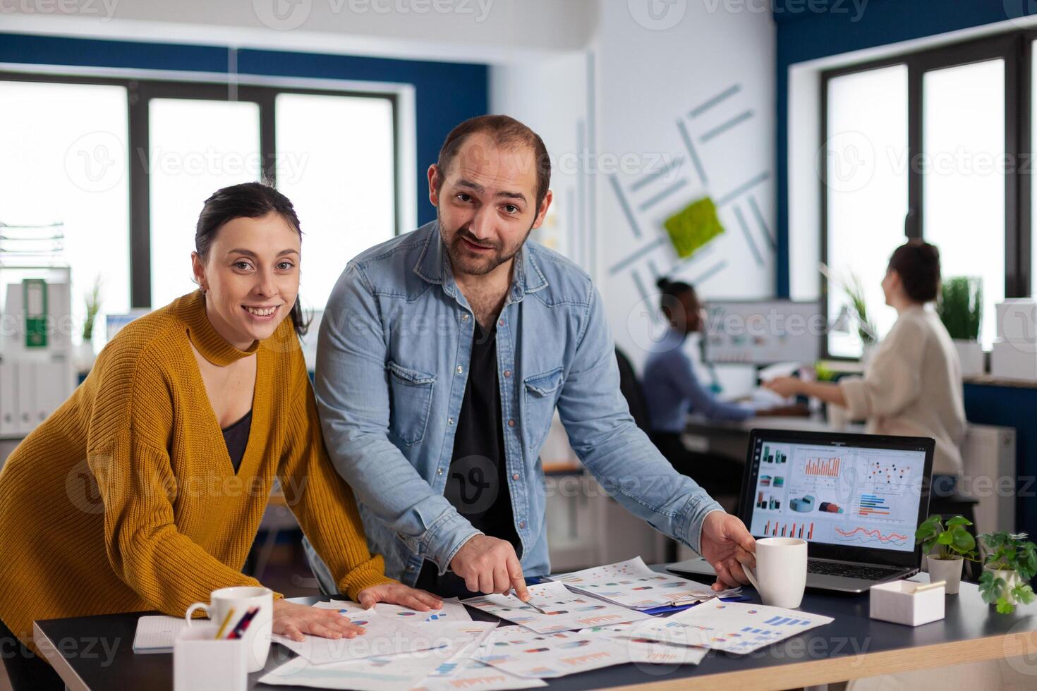 Multiethnic management smiling at camera working to finish project pointing at charts on desk. Diverse team of business people analyzing company financial reports from computer. photo