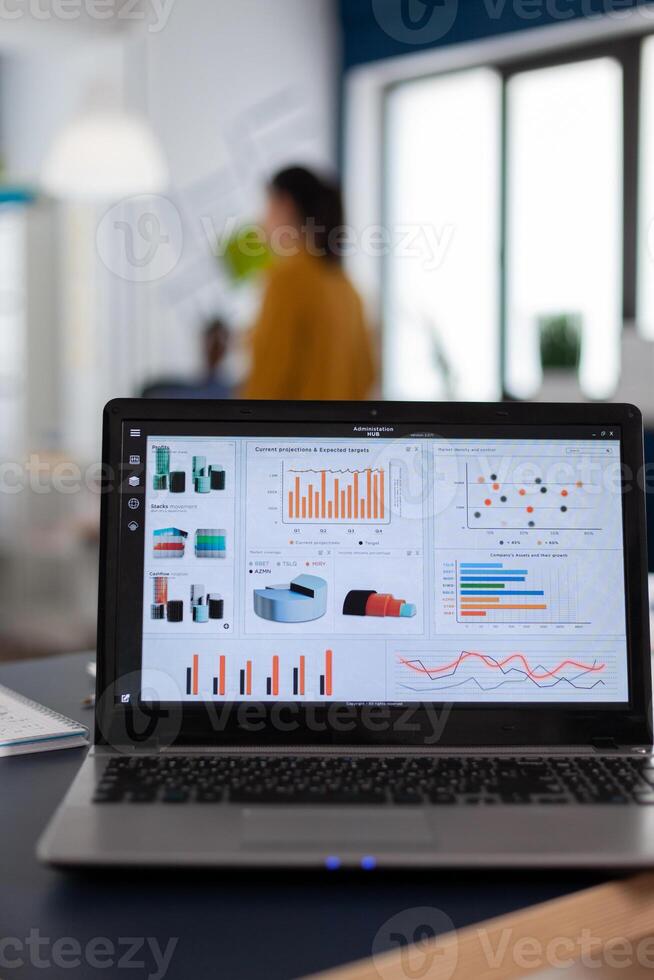 Laptop with marketing charts, graphs analitics in start up company. Workplace area in business center with multi ethnic employees in it, shot of room with modern furniture and blue wall. photo