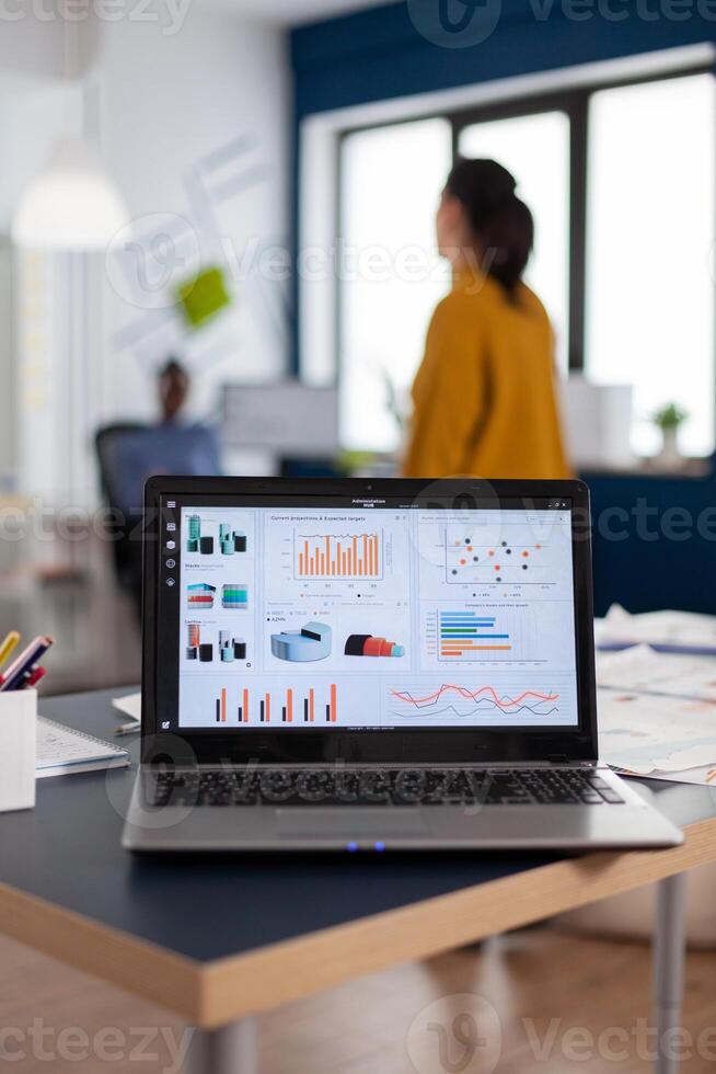 Close up of laptop with graphs, charts of marketing agency. Workplace area in business center with multi ethnic employees in it, shot of room with modern furniture and blue wall. photo