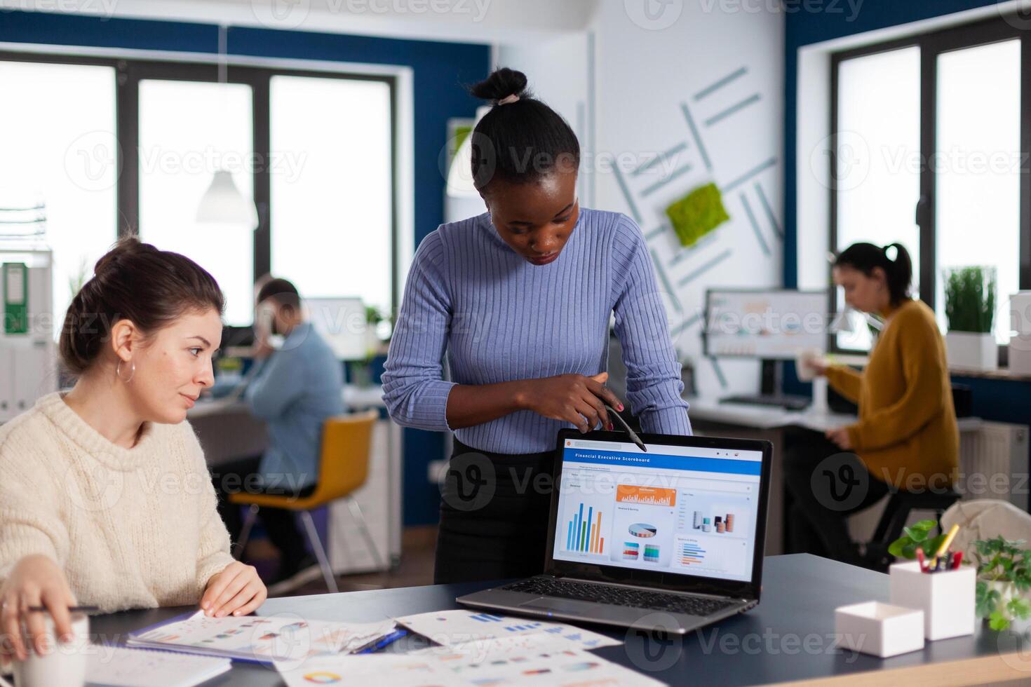 African manager of startup company briefing employee about new financial marketing project pointing at charts. Multiethnic employees gathered in co-working space. photo