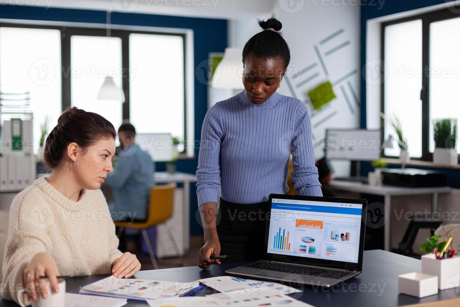 Diverse businesswomen discussing new project for company evolution, black woman checking tasks on new contract. Multiethnic employees gathered in co-working space. photo