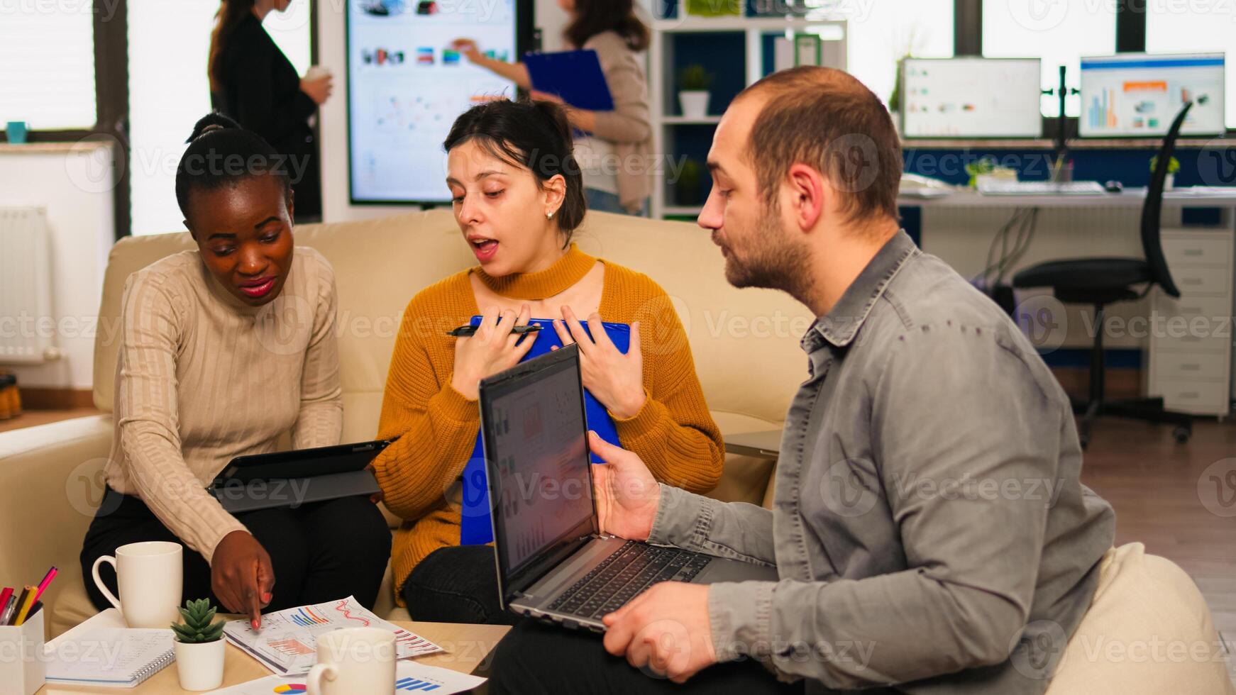 Confident company manager giving working tasks to diverse teammates sitting on couch in start up office. Multiethnic team discussing project ideas at brainstorming meeting using digital devices photo