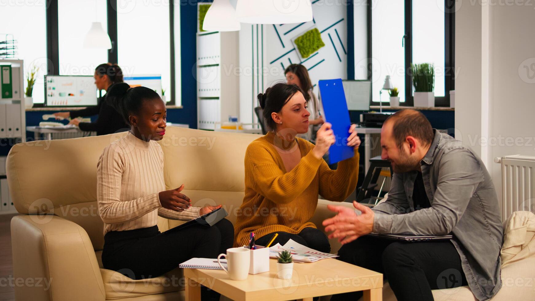 Angry manager woman screaming at diverse employees disagreeing about bad business contract, hitting man with clipboard in head sitting on couch at office desk, conflict dispute about document photo