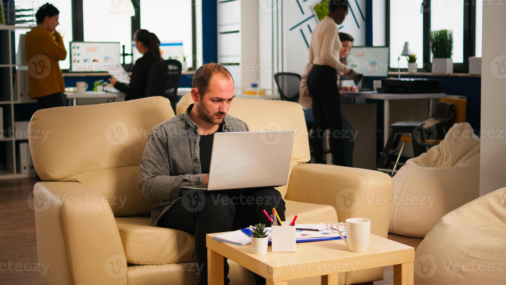 Business man typing on laptop sitting on couch in start up office while diverse team working in background analysing statistics data. Multiethnic coworkers talking about startup financial company photo
