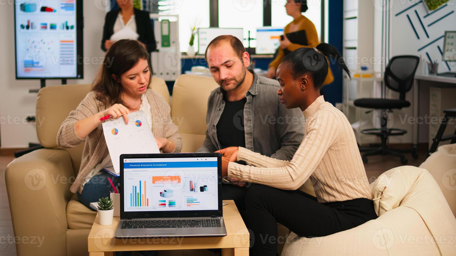 Diverse colleagues employees cooperating in office discussing at workplace, analysing graphs sitting on couch. Business coworkers planning start up business project, brainstorming in corporate office photo