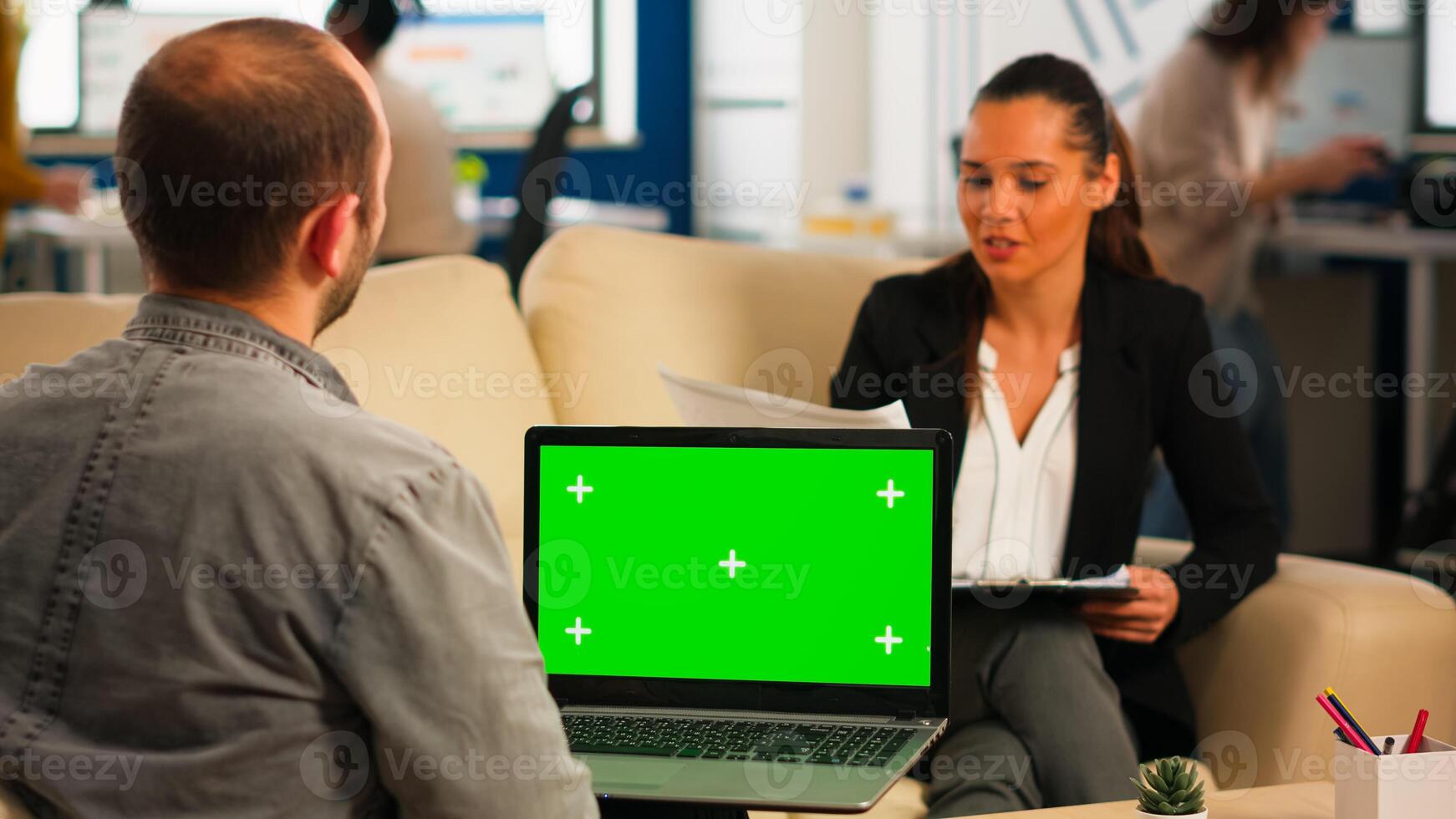 Over the sholder of manager sitting on couch talking with colleague holding laptop with green screen while diverse team working on background. Multiethnic people planning project on chroma key display photo