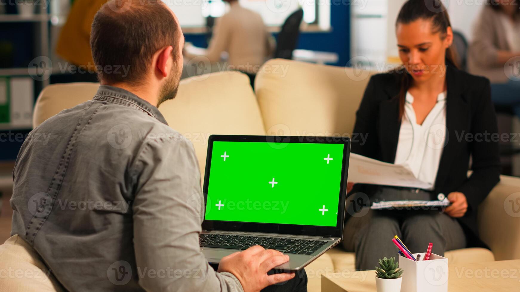 Back view of business man sitting on couch using laptop with green screen talking with colleague while team working on background. Multiethnic coworkers planning project on chroma key display photo