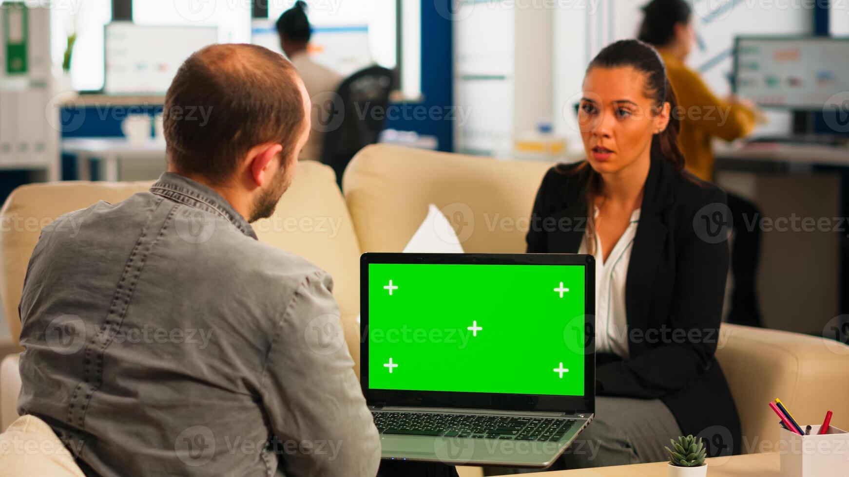 Business team sitting on couch analysing financial statistics, holding laptop with green screen while diverse team working on background. Multiethnic coworkers planning project on chroma key display photo