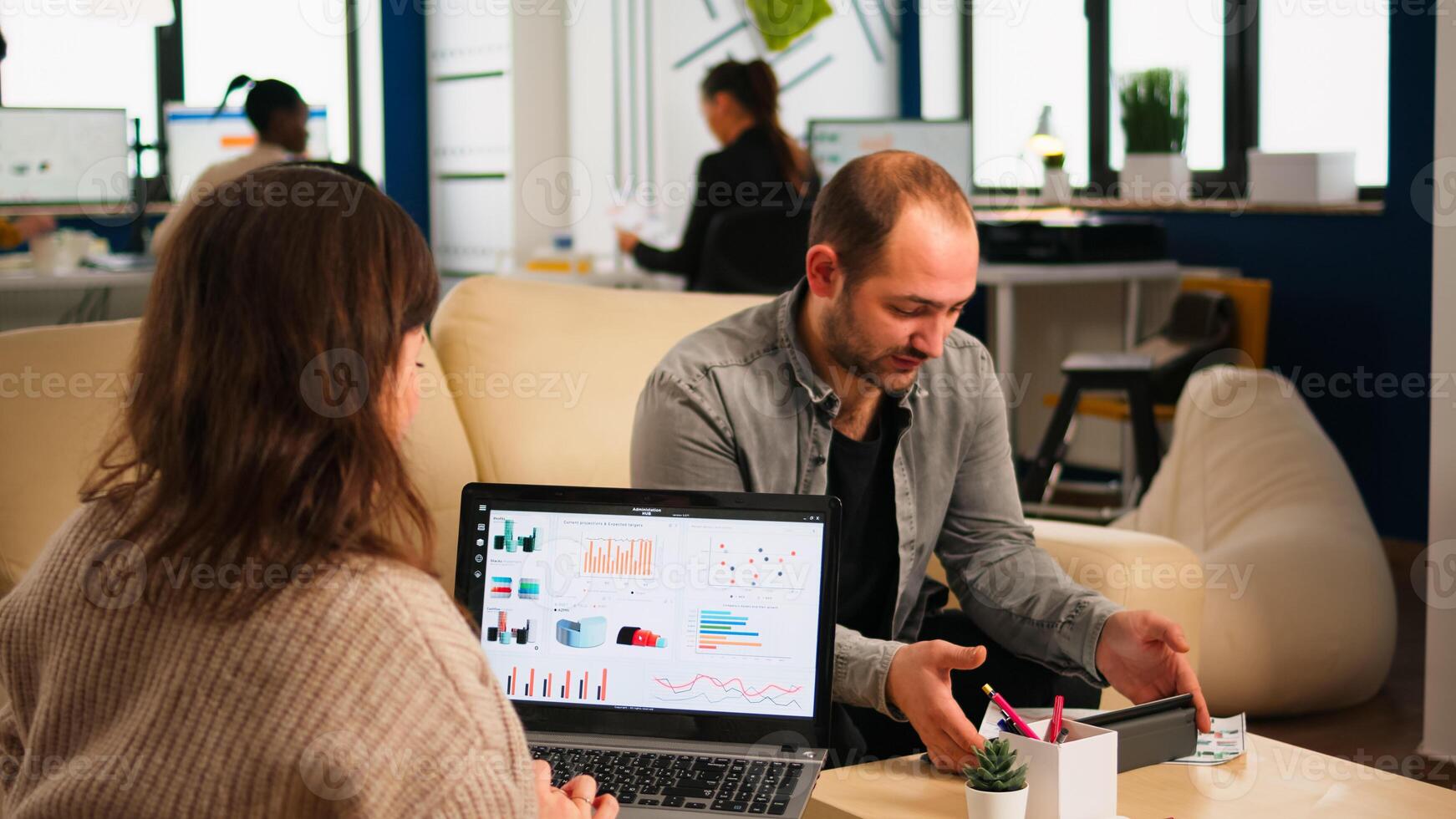 Close up of back view young entrepreneur sitting on couch in office using laptop computer while colleagues working on background. Multiethnic coworkers planning new financial project in modern company photo