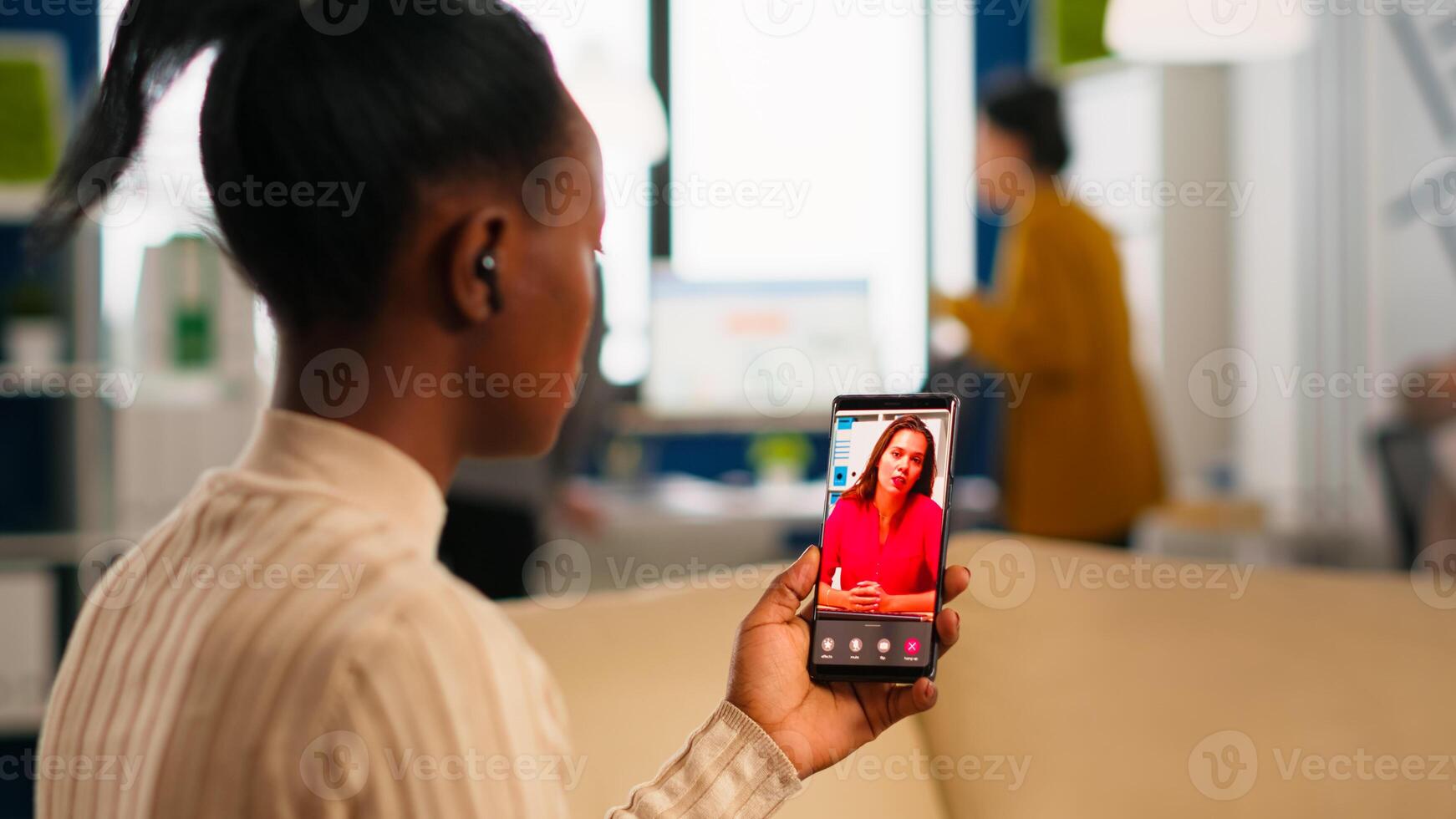 Back view of african woman discussing with remote manager on call holding smartphone using headphones sitting on couch in business modern office. Diverse coworkers planning new financial project photo