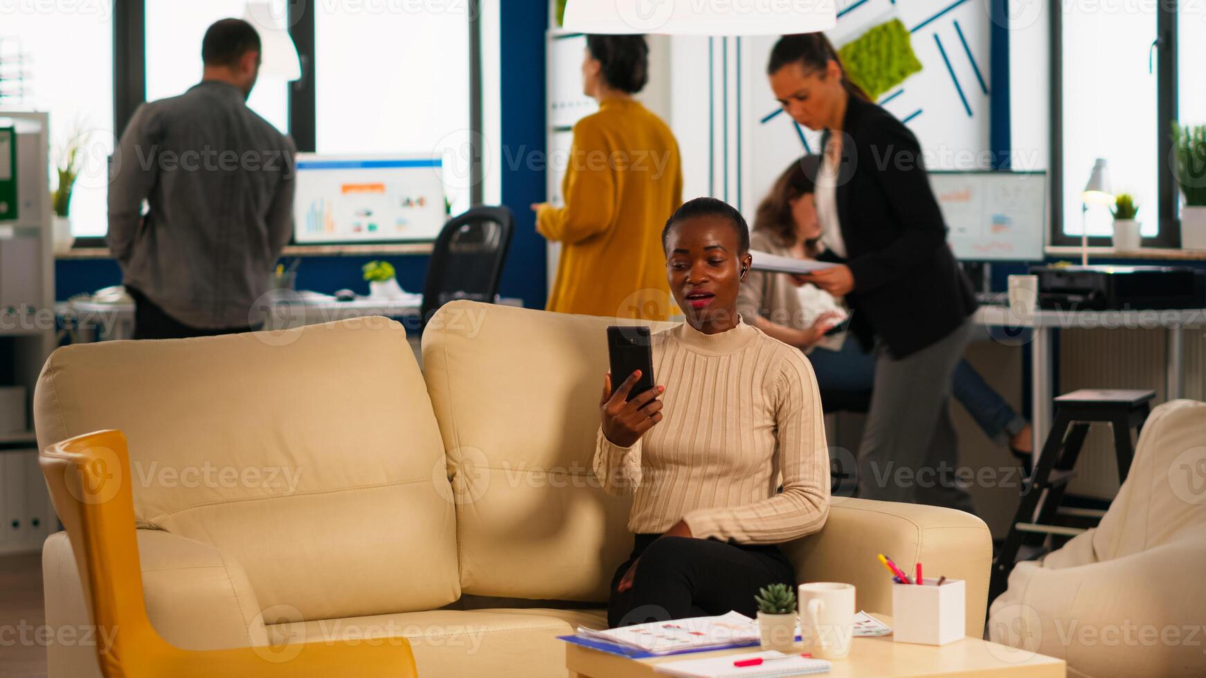 Tired black woman talking with colleague using call holding smartphone with wiraless headset sitting on couch in business office. Diverse team planning new financial project in modern company photo