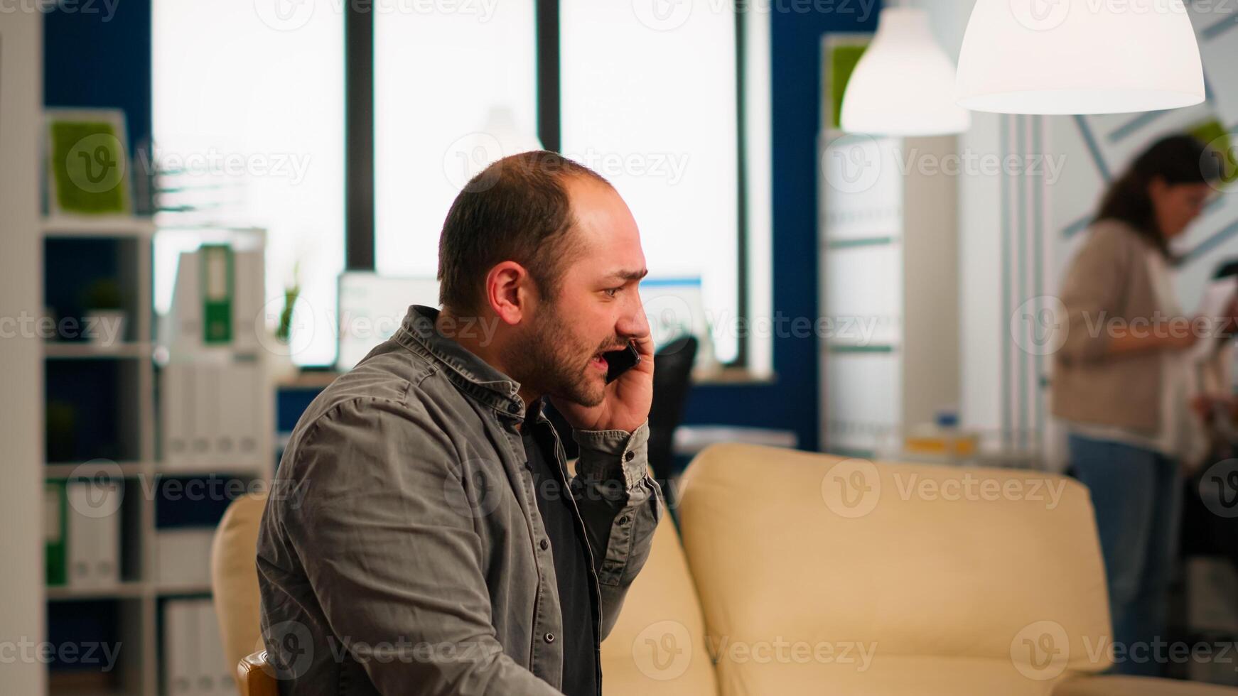 Nervous manager aggressively talking on phone and curses, screaming sitting on chair in middle of business office. Multiethnic scared coworkers planning financial project working in modern company photo