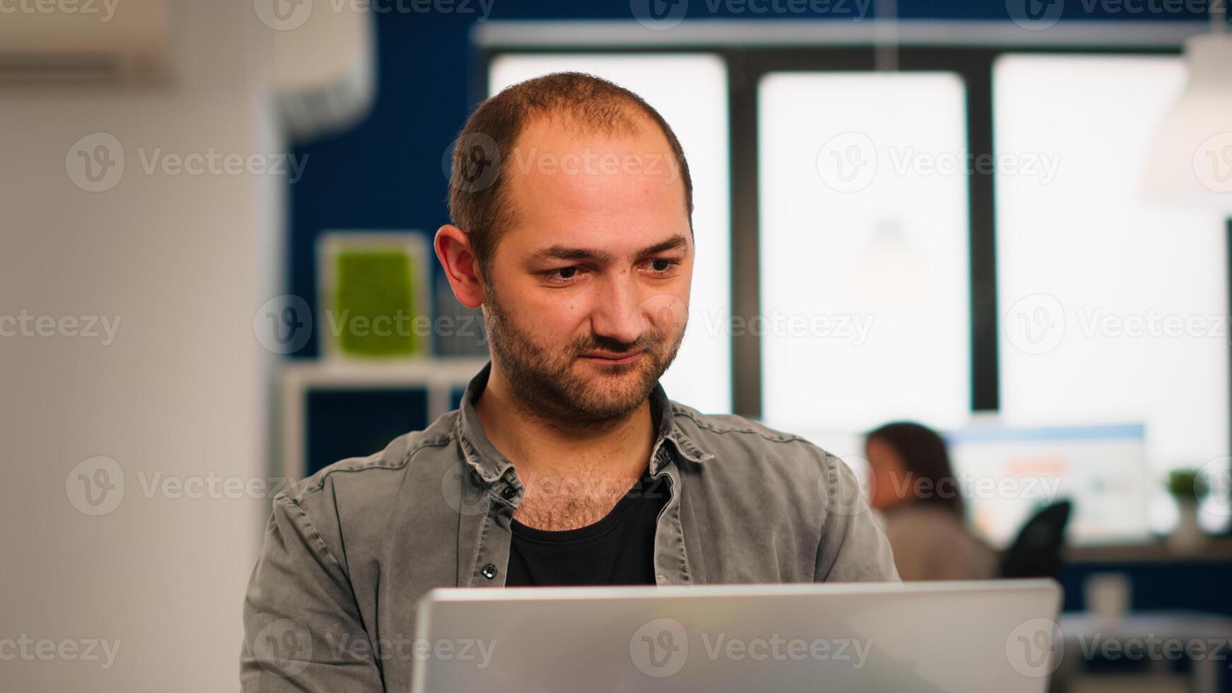 Enthusiastic business man writing a report sitting at table in modern startup business office sending mails typing on laptop, greeting colleague. Man working in financial company with diverse team. photo