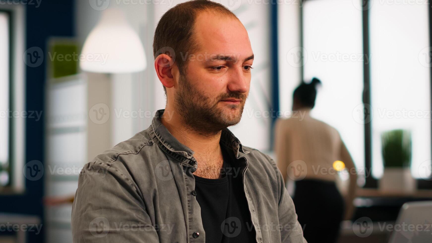 Close up of authentic manager sitting at table in modern startup business office sending mails typing on laptop smiling and greeting colleague. Man working in financial company with diverse team. photo