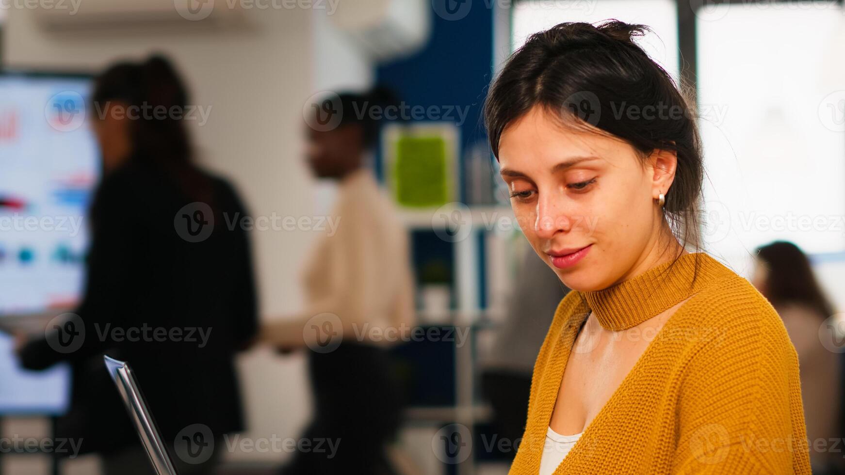 Young woman in casual clothing working at laptop sitting at desk in busy financial startup company while diverse team analyzes statistics data in modern office. Multiethnic team concentrated on work photo