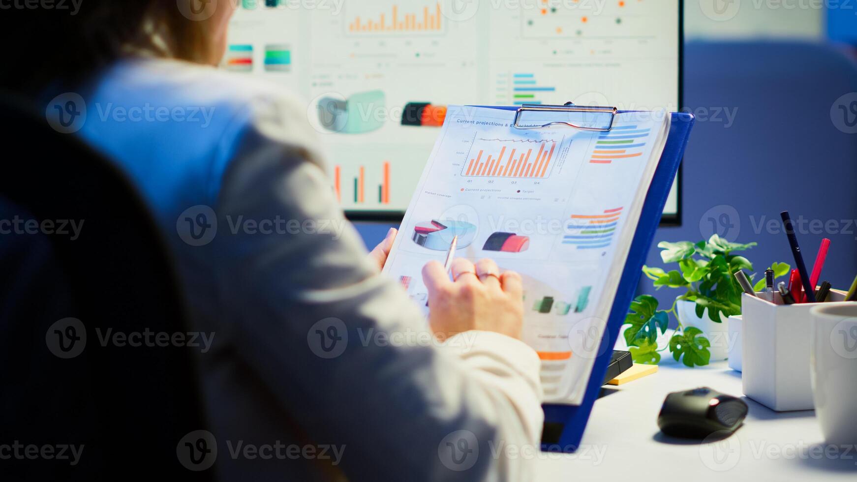 Close up of business woman holding clipboard with graphics and financial statistics working overtime in front of computer sitting in start-up business office. Busy employee using modern technology photo