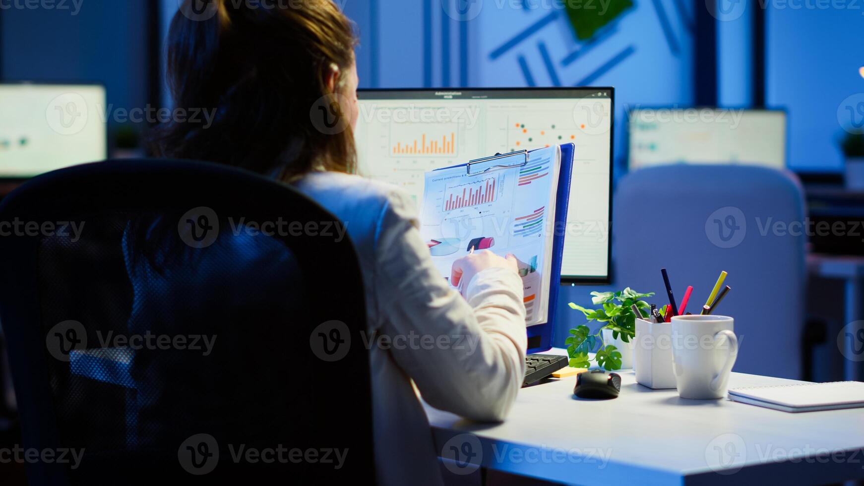 Back view of manager analysing graphics and financial statistics from clipboard working overtime in front of computer sitting in start-up business office. Busy focused employee using modern technology photo